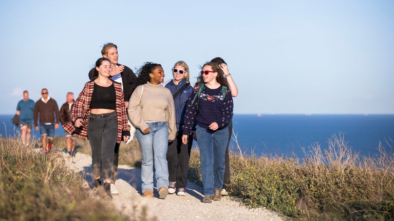 A group of young people walk along a coastal path towards the camera with the sea visible behind them